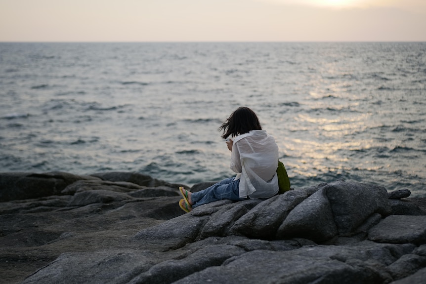 A woman sits on a rocky beach and looks at her phone