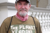 Chris Hooper looking at the camera, straight face, facial hair, blue eyes, wearing a cap, t-shirt says "the planet vs adani".
