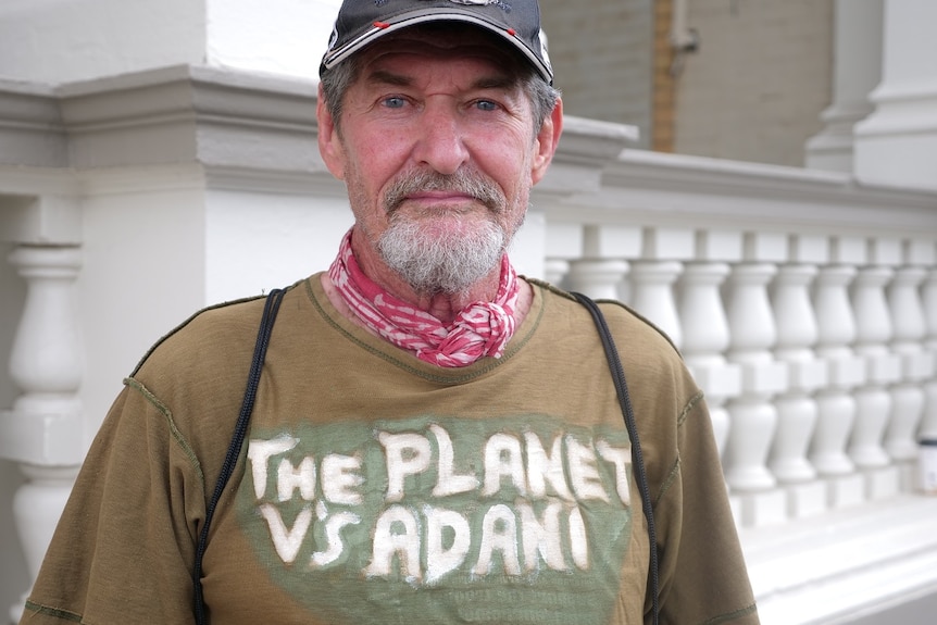Chris Hooper looking at the camera, straight face, facial hair, blue eyes, wearing a cap, t-shirt says "the planet vs adani".