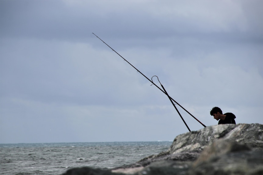 Grey skies seen over the ocean, as a man fishes off rocks in the foreground. 