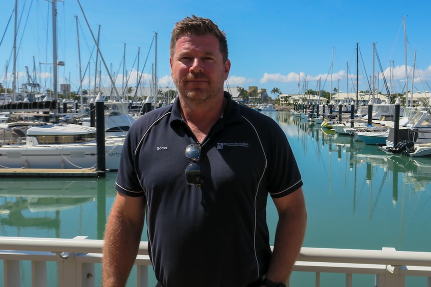 A man looks at the camera. He is standing at a marina.