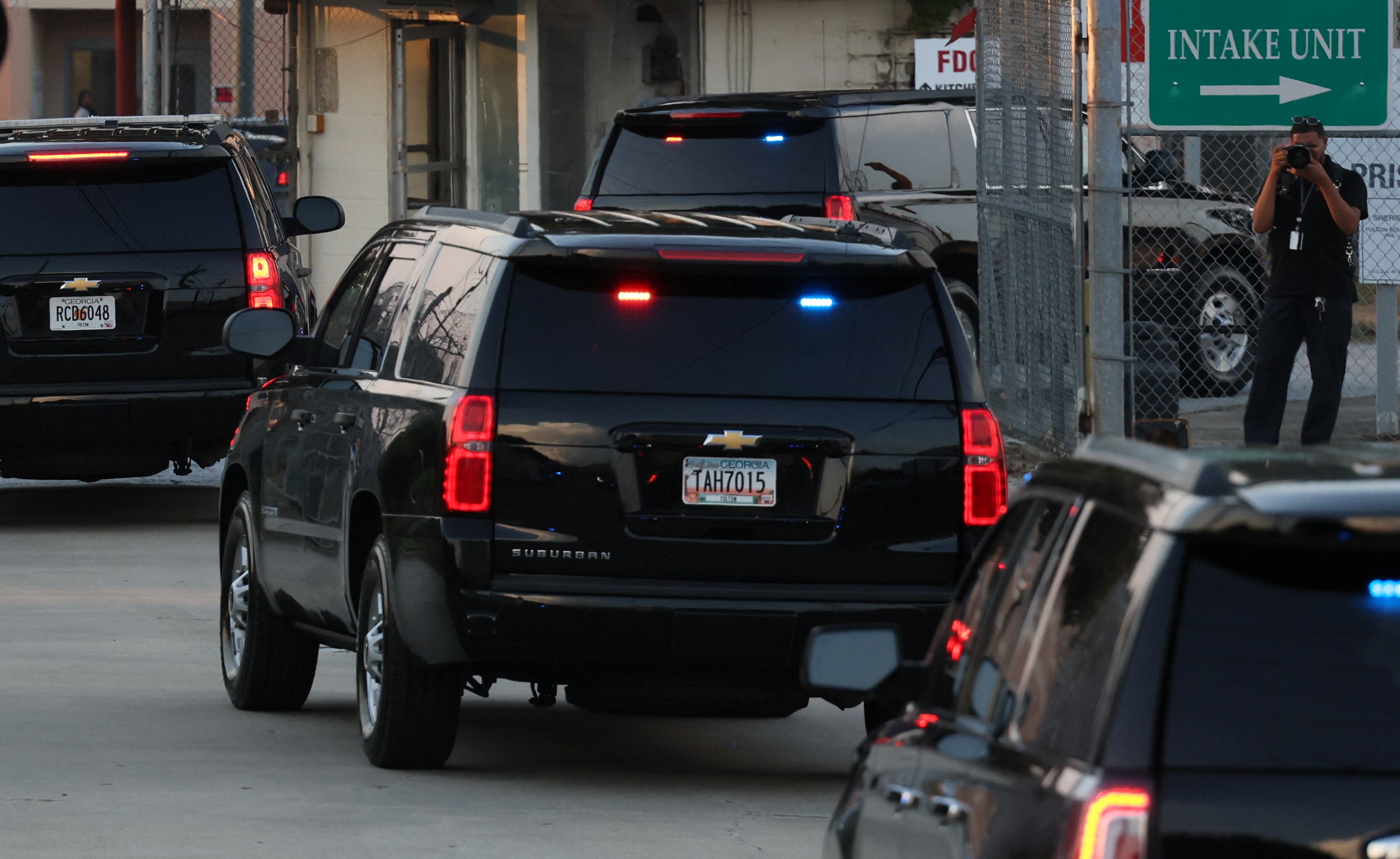 A black car carrying Donald Trump drives behind several others, through a gate with a sign that says INTAKE UNIT
