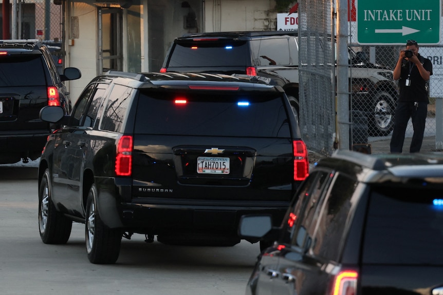 A black car carrying Donald Trump drives behind several others, through a gate with a sign that says INTAKE UNIT