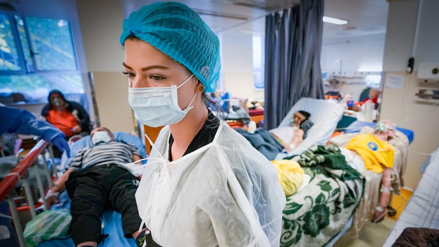 A female health worker in a mask stands amidst several hospital beds holding COVID patients