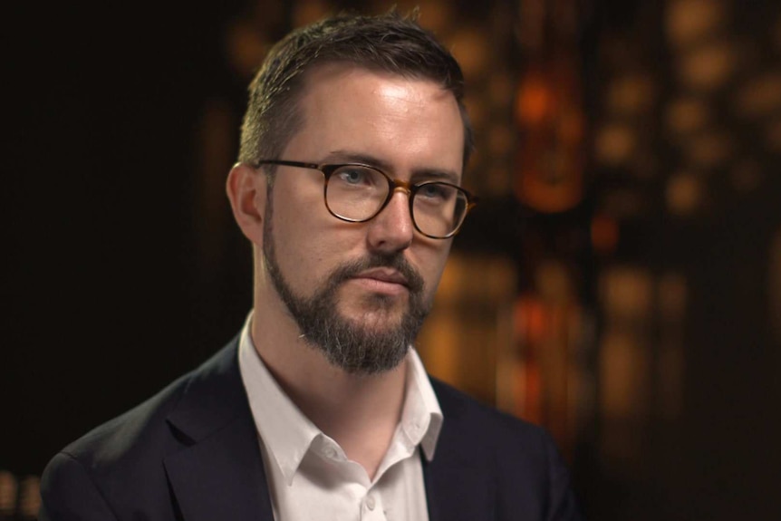 Head shot of Fergus Ryan wearing open-collared white shirt, grey suit and glasses