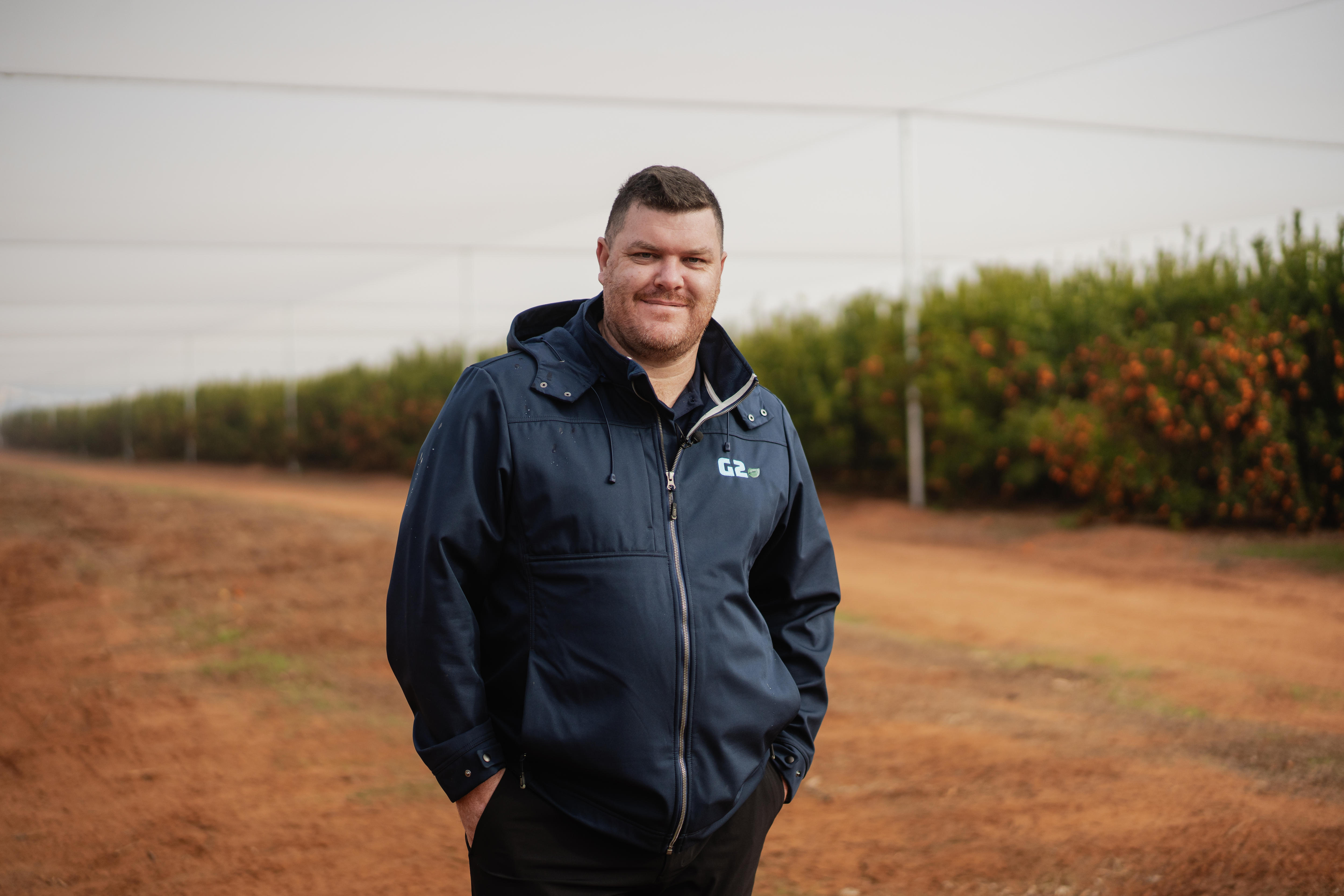 Un hombre sonríe a la cámara frente a unos naranjos.
