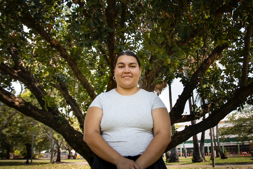 A young woman wearing a white shirt smiles at the camera. Behind her is a large tree.