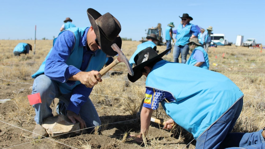 Outback Queensland grazier kneels in dirt, alongside son, using pick to dig for fossils.