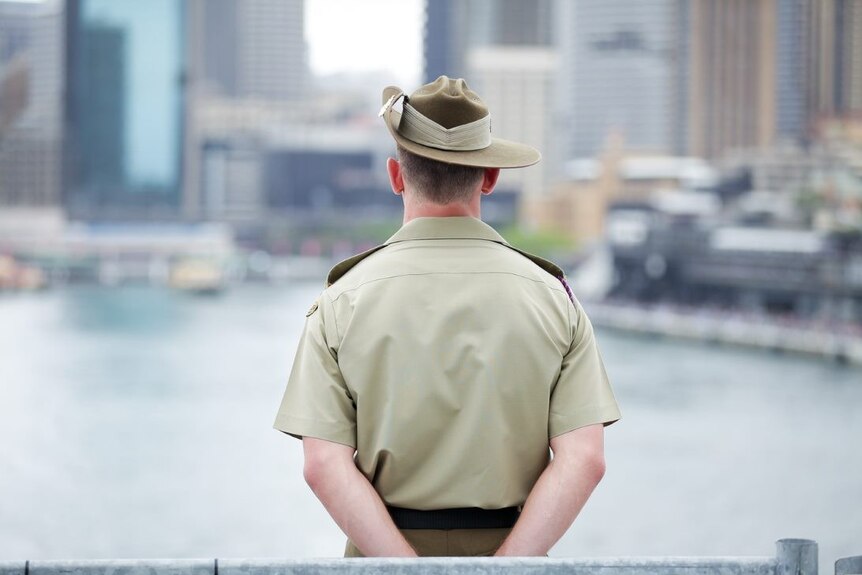 A military officer stands with his back to the camera and overlooking a river with high-rise buildings in the background.