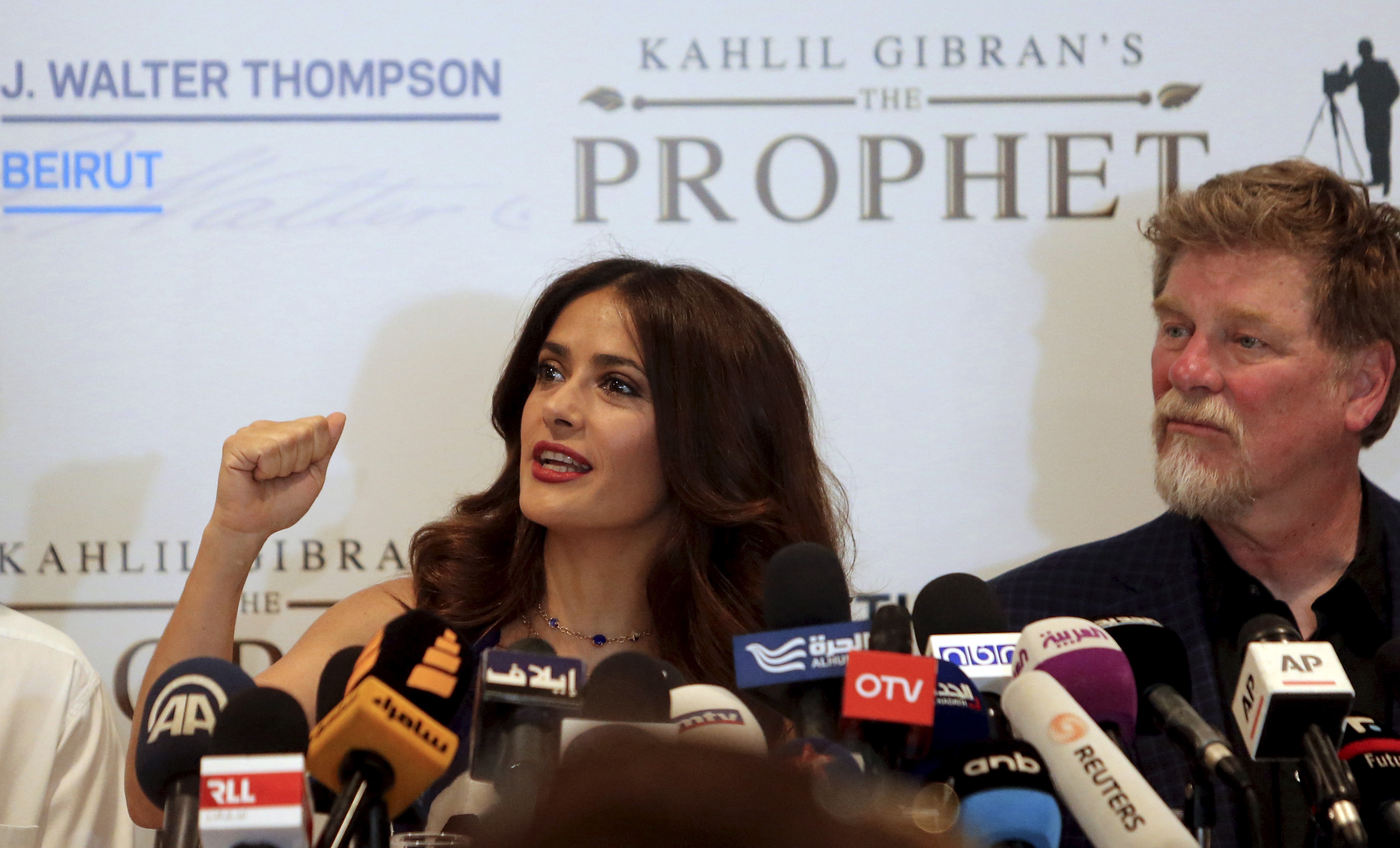 Salma Hayek sentada ao lado de Roger Allers (à direita) com vários microfones à sua frente.