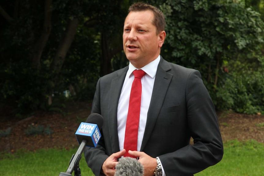 A man in a dark suit and a bright tie stands outside speaking into a microphone.