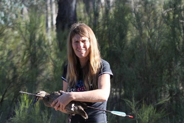 Woman with long hair stands in bushland holding an injured duck that's been shot by an arrow