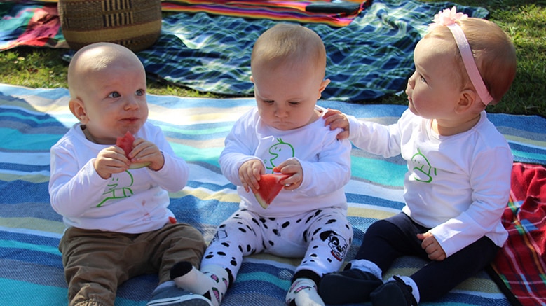 Three babies sitting on a mat together eating watermelon.