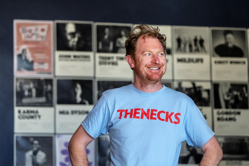Man in blue t-shirt that has The Necks written in red lettering stands in front of wall of posters advertising live music 