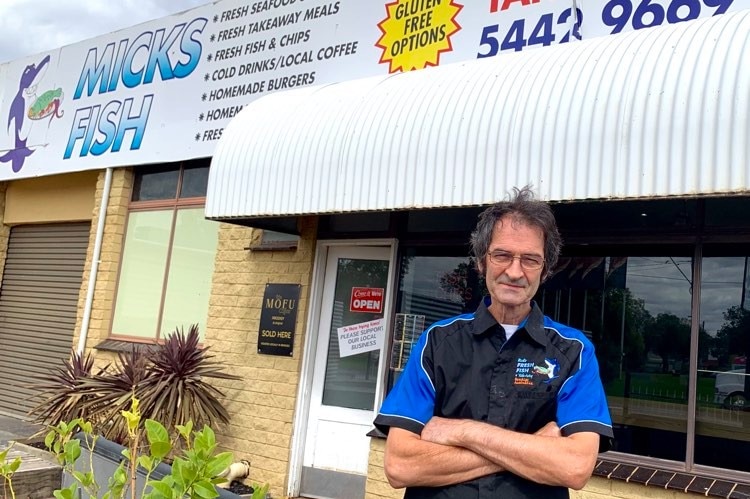 A man is standing outside a fish and chip shop folding his arms, wearing glasses looking at the camera.