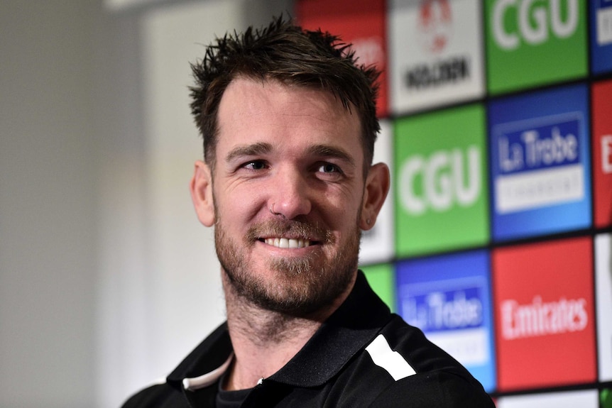 Dane Swan smiles in front of a wall of sponsor logos.