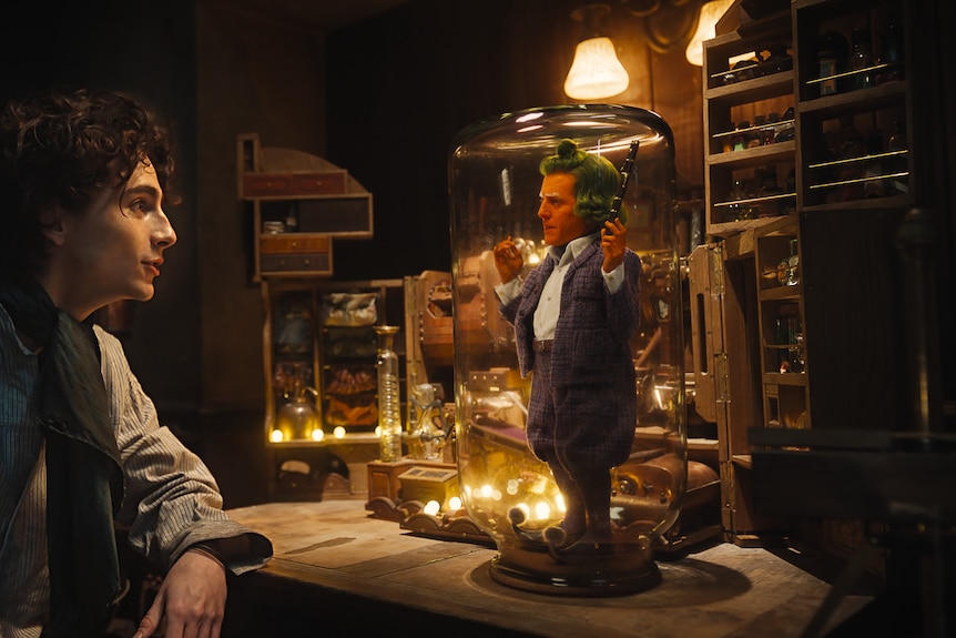 Wonka talks to a small orange man played by Hugh Grant, with green hair, standing under a glass jar.