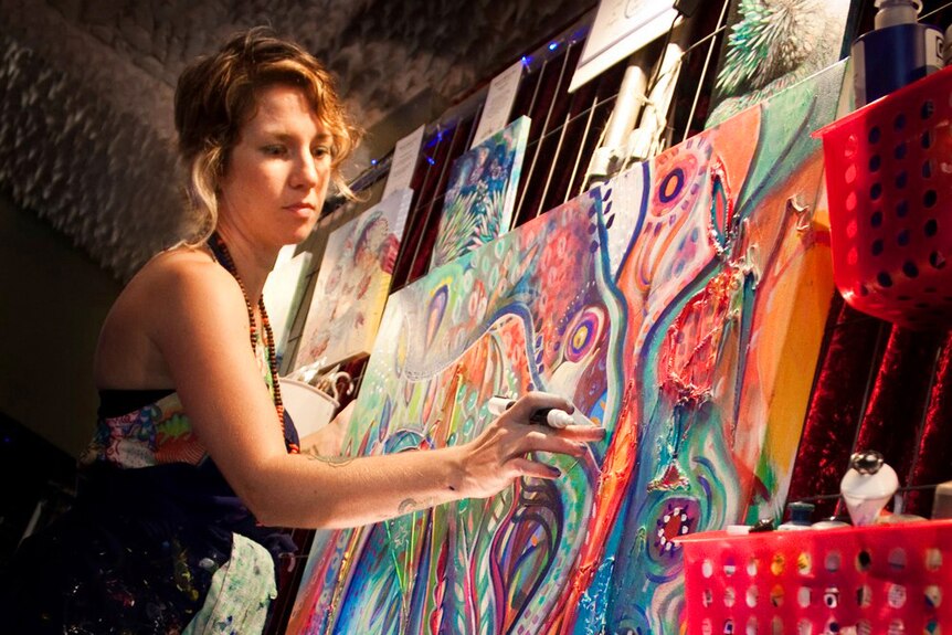 A young woman with short hombre hair wears an artist's apron and is painting a large colourful canvas.