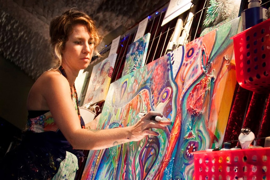 A young woman with short hombre hair wears an artist's apron and is painting a large colourful canvas.