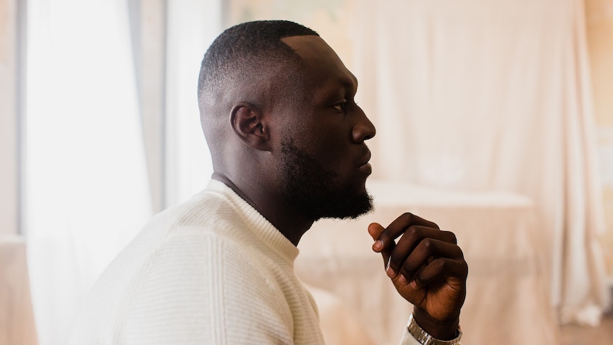 A side on profile of Stormzy wearing a white knit sweater and lifting his hand to his chin