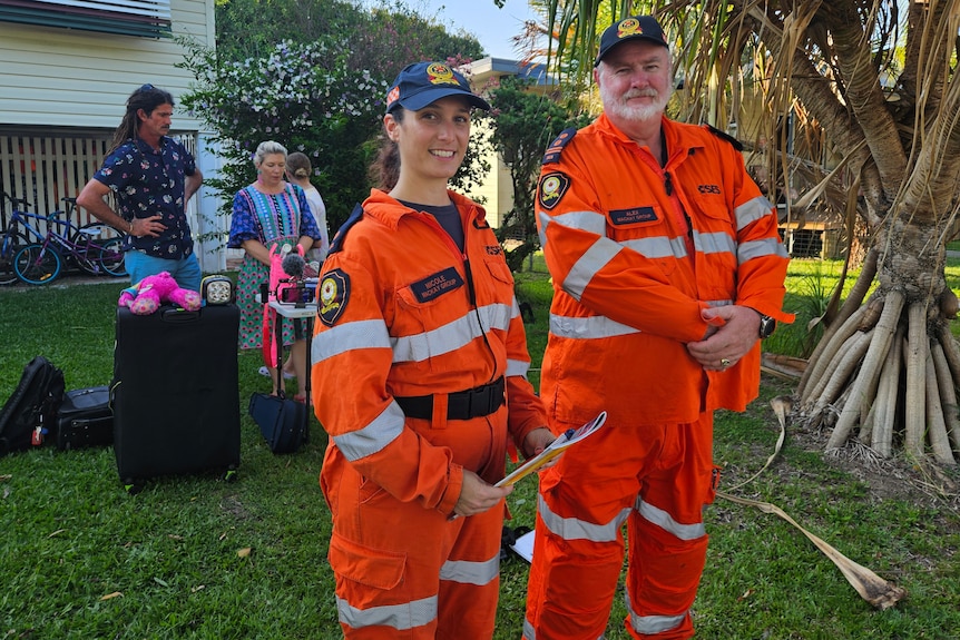 A man and woman in SES uniforms smiling at the camera with a family in the background.
