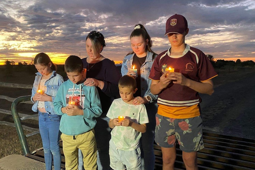 A woman and her five children stand on a driveway at sunset looking solemn and each holding a candle