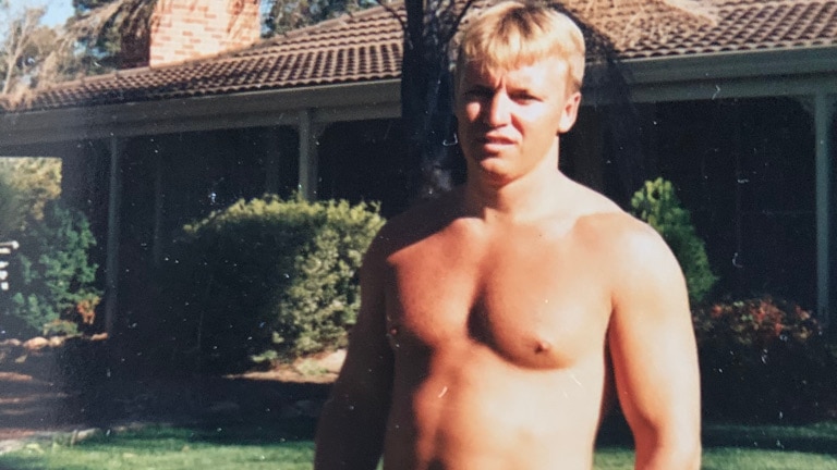 A 1997 photograph of a blonde man with no shirt on standing outside a house on the grass