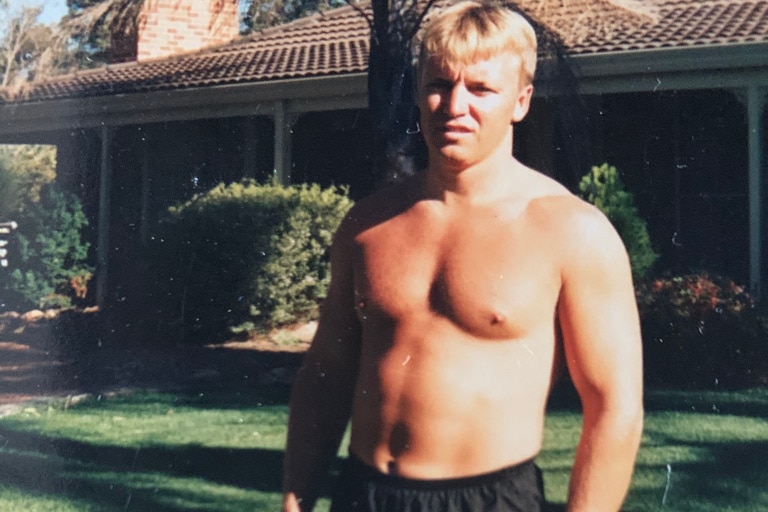 A 1997 photograph of a blonde man with no shirt on standing outside a house on the grass