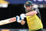 A man in a green helmet and yellow and green Australian cricket uniform plays a shot with a Gray-Nicolls cricket bat.