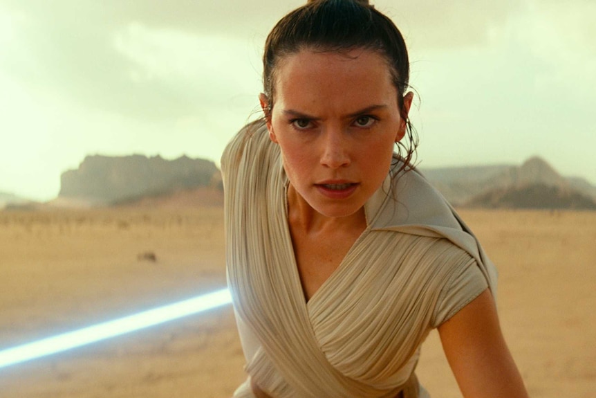 A woman wearing a white robe and carrying a lightsaber stands in a desert.