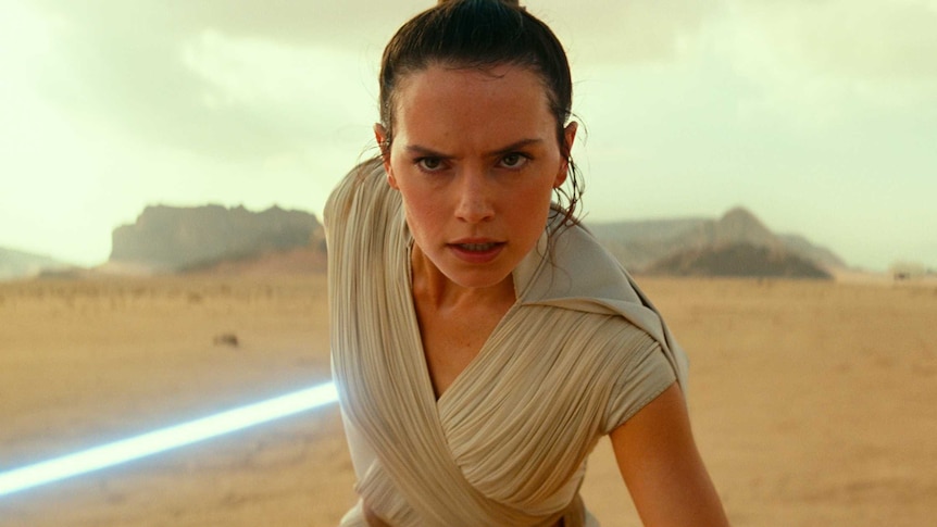 A woman wearing a white robe and carrying a lightsaber stands in a desert.