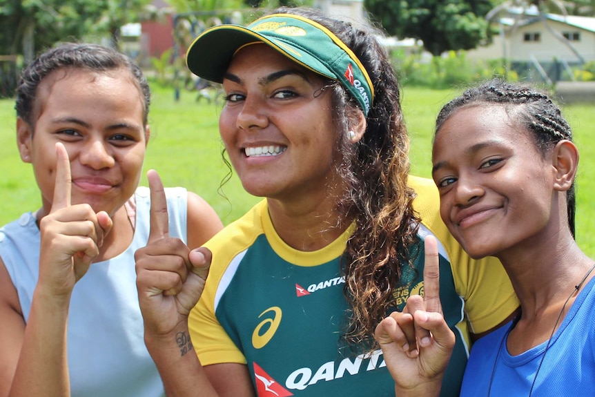 An Australian rugby player is flanked by two Fijian girls. They all make the Number 1 signal with their hands.