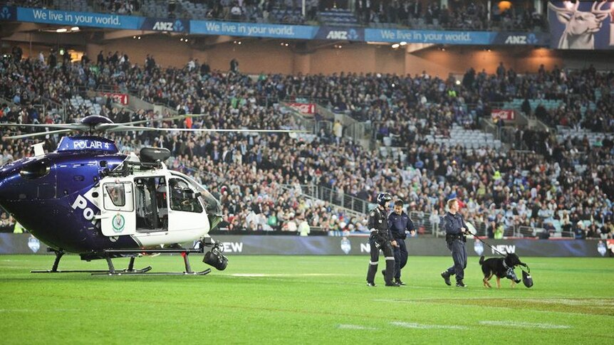 A police helicopter in ANZ stadium with three police officers walking Chuck who is carrying the ball.