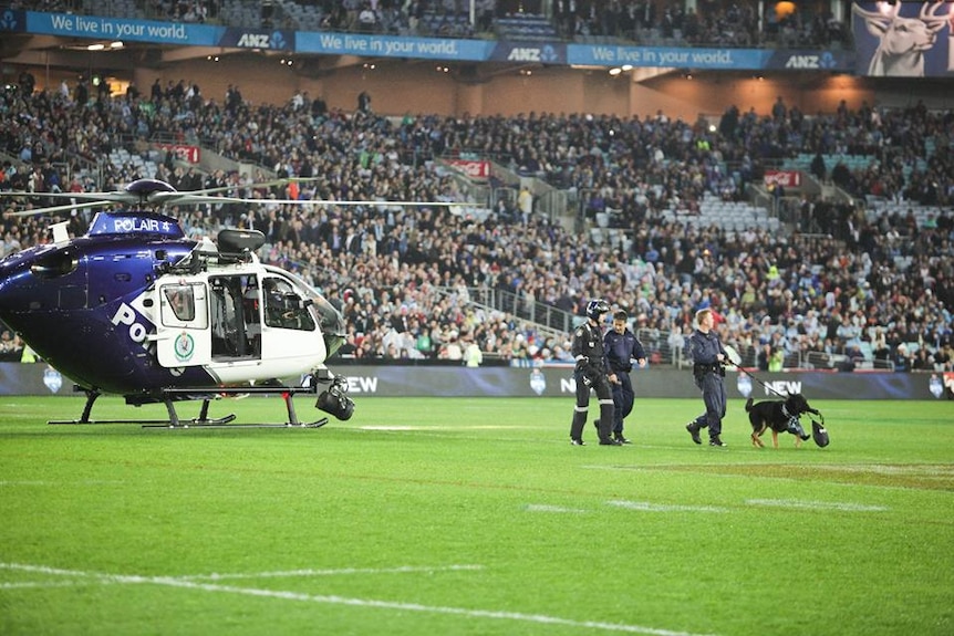 A police helicopter in ANZ stadium with three police officers walking Chuck who is carrying the ball.