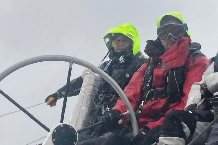 Two men in rain coats sit behind a yacht steering wheel