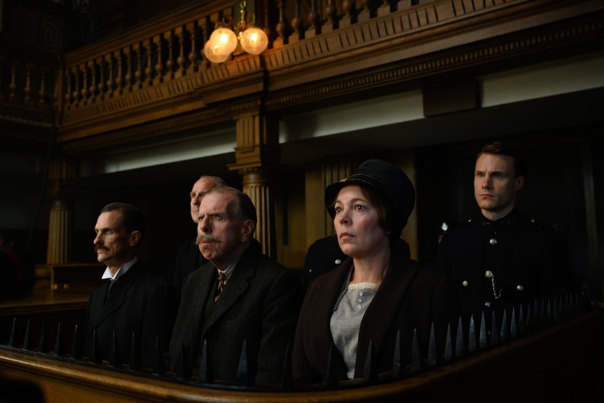 Two rows of people dressed in 1920s attire stand in the gallery of a courtroom.