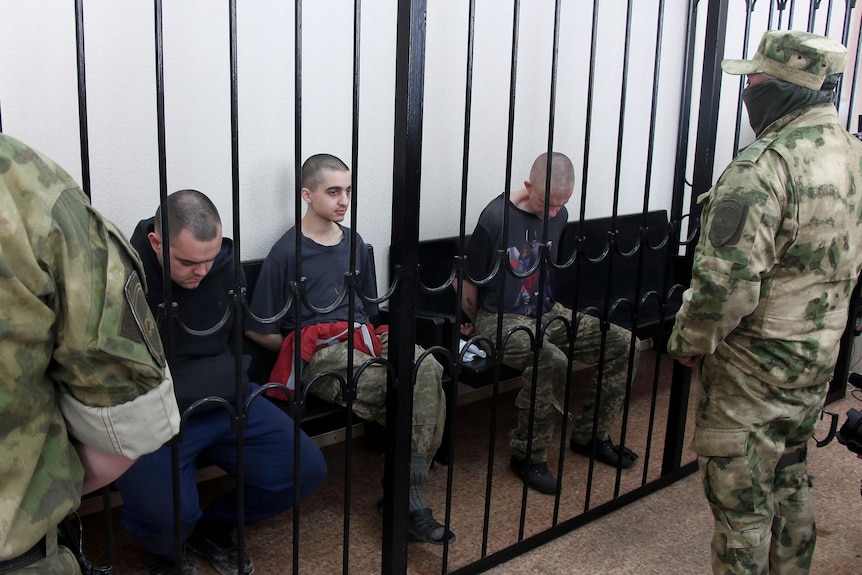 Three men sit on a bench inside a cell as two masked soldiers stand guard.