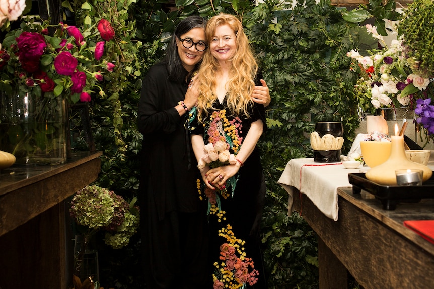 Two women stand closely surrounded by lucious foliage and flowers, both smiling