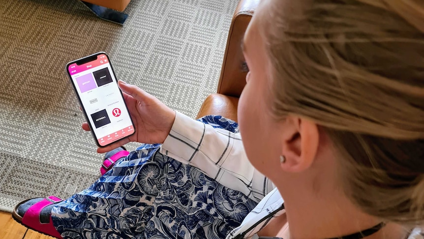 Looking over the shoulder of a woman perched on a couch holding a phone with a shopping app open.