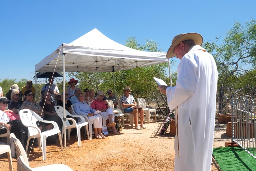 A bishop leading a service in the bush with a small crowd of people seated in front of him.