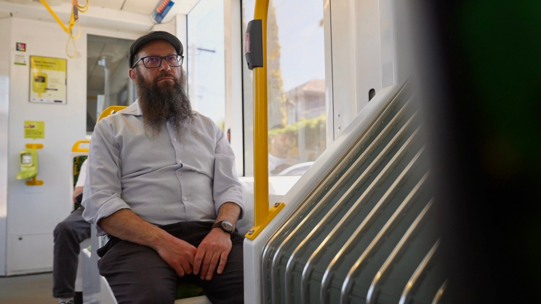Um homem com uma longa barba grisalha, óculos e um boné cinza com uma camisa branca de botões está sentado em um bonde, olhando para frente.