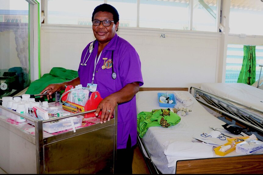 Woman in bright purple nurse uniform shirt stands next medical supplies in a hospital room. 