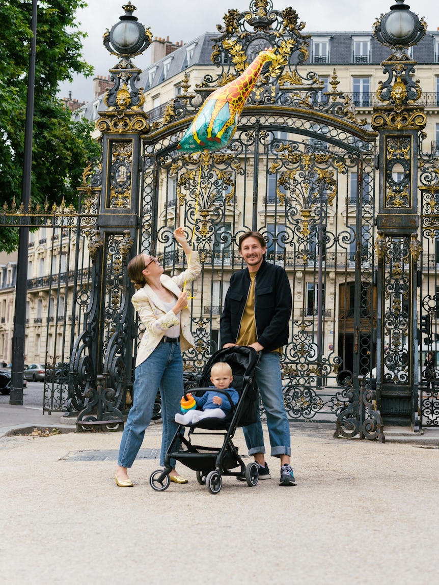 Ένας άντρας και μια γυναίκα στέκονται με τον μικρό σας γιο σε ένα καροτσάκι ανάμεσά τους και ένα κτίριο στο Παρίσι πίσω τους.