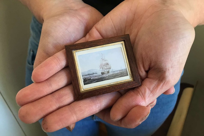 Close up of open palmed hands holding a tiny painting of a convict ship