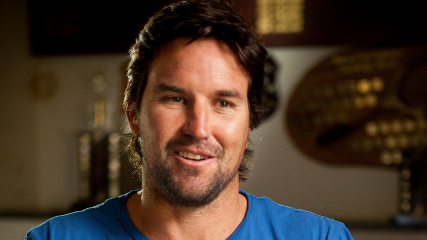 A brown-haired man with stubble sits down while wearing a plain blue T-shirt, with sporting trophies in the background.