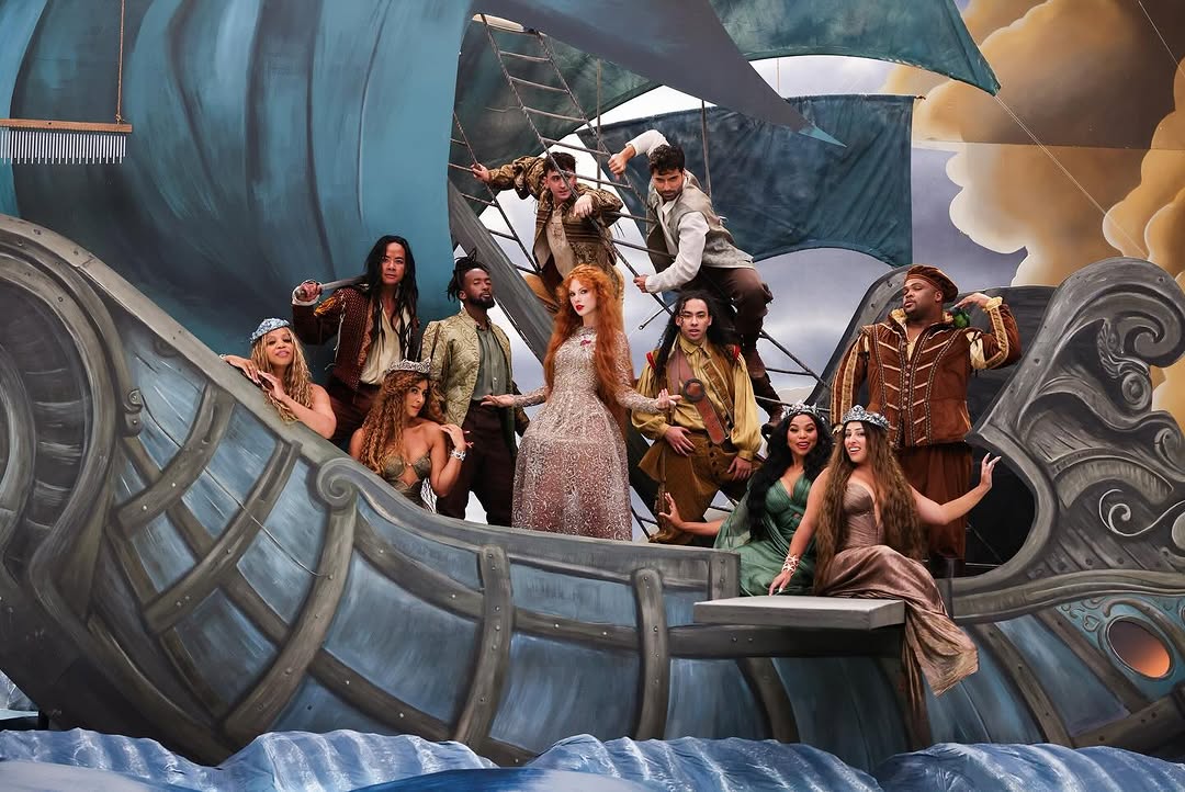 A woman stands wearing a silver dress on a play set of a boat in an ocean with people onboard