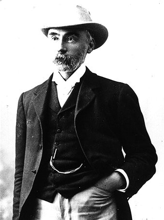An old black and white photo of a man in a smart hat, black jacket and a beard.