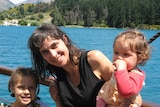 A smiling dark-haired woman holds two children in front of a scenic lake in New Zealand.