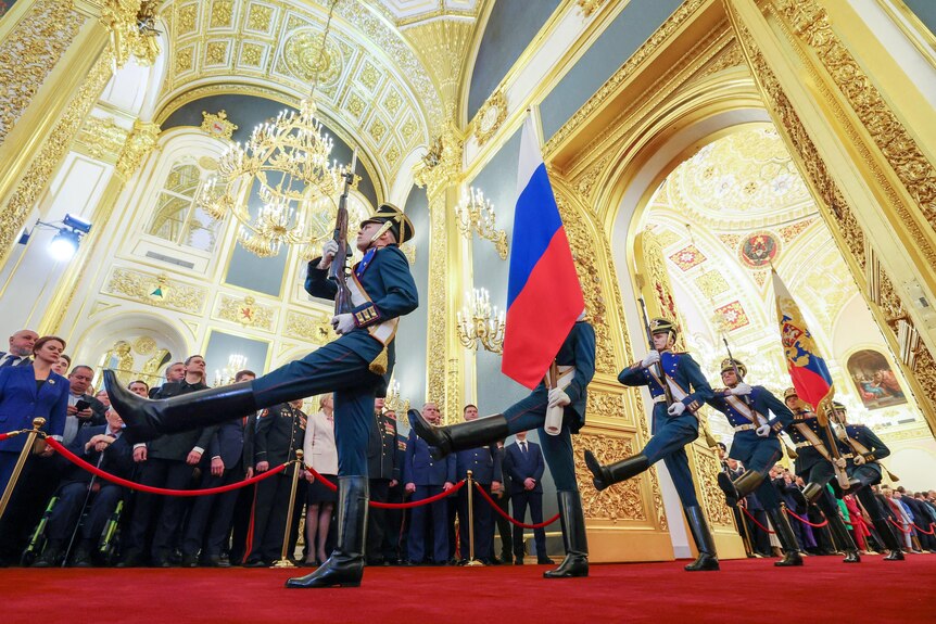 Honour guards carry guns and Russian flags through a gold-painted hall with red carpet as people watch on