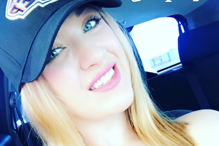 A woman wearing a hat and black singlet top takes a selfie while sitting in a car.